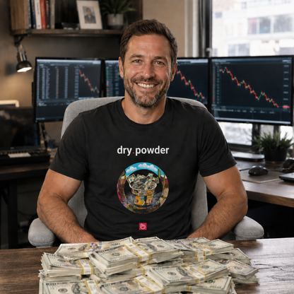 Smiling 37-year-old trader sitting in a bright home trading office wearing a black dry.powder nano.narrative T-shirt. Red declining charts appear on multiple monitors behind him, while stacks of $100 bills are spread across the desk without covering the shirt design. The trader sits on a light gray chair and looks directly at the camera in natural daylight.